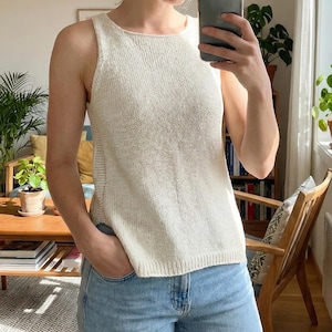 May include: Cream-colored knitted tank top with a round neckline and a sleeveless design. The top is made of a textured knit fabric. The person is wearing blue jeans. The background includes a wooden chair, a coffee table, and plants.