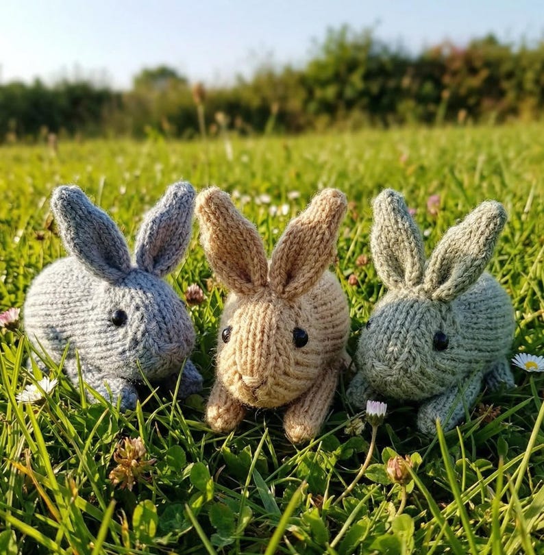May include: Three knitted bunny rabbits in shades of gray, tan, and green sit in a field of green grass and small white flowers. The bunnies have black button eyes and long ears. The scene is bathed in sunlight.