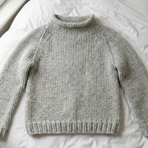 K&ouml;nnte beinhalten: Ein hellgrauer Strickpullover mit hohem, geripptem Kragen und langen &Auml;rmeln. Der Pullover hat einen gerippten Saum und B&uuml;ndchen. Das Strickgewebe hat ein gesprenkeltes Aussehen. Der Pullover liegt auf einer wei&szlig;en Oberfl&auml;che.