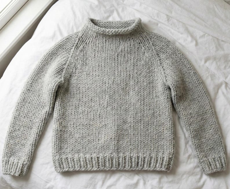 K&ouml;nnte beinhalten: Ein hellgrauer Strickpullover mit hohem, geripptem Kragen und langen &Auml;rmeln. Der Pullover hat einen gerippten Saum und B&uuml;ndchen. Das Strickgewebe hat ein gesprenkeltes Aussehen. Der Pullover liegt auf einer wei&szlig;en Oberfl&auml;che.