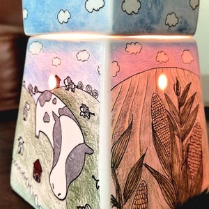 K&ouml;nnte beinhalten: Ein wei&szlig;er Keramik-Wachsschmelzer mit Bauernhof-Motiv. Der Schmelzer zeigt einen blauen Himmel mit wei&szlig;en Wolken oben, eine Kuh auf einem Feld mit einem roten Stall und Maispflanzen an der Seite.
