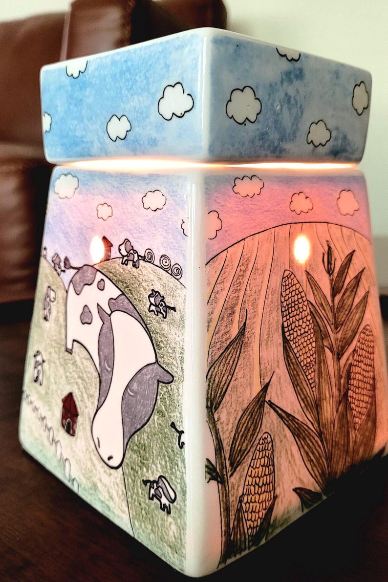 K&ouml;nnte beinhalten: Ein wei&szlig;er Keramik-Wachsschmelzer mit Bauernhof-Motiv. Der Schmelzer zeigt einen blauen Himmel mit wei&szlig;en Wolken oben, eine Kuh auf einem Feld mit einem roten Stall und Maispflanzen an der Seite.