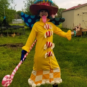May include: A whimsical costume featuring a large, striped hat with lollipops, a yellow dress adorned with candy accents, and a matching candy cane staff. The outfit includes a red hat, blue polka-dot wings, and a checkered skirt.