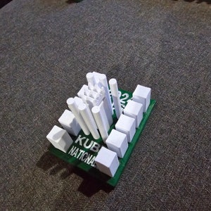 May include: A white 3D printed model of a city skyline on a green base with the text "KUB NATIONAL" printed on it.