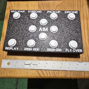 May include: A black control panel with white buttons and white text. The text reads "Mulligan Scorecard", "Pickup Drone", "Aim", "Replay", "Green View", "Green Grid", and "Fly Over".