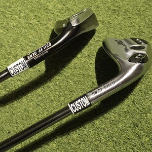 May include: Two golf clubs with black shafts and silver club heads are positioned diagonally against a green background. Each club has a white label that reads "CUSTOM". The club heads have text that is difficult to read.