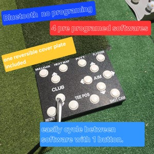 May include: Black golf training device with white buttons labelled "MULLIGAN", "HEAT MAP", "PUTT", "FLYOVER", "CLUB", "AIM", "TEE POS", and "SHOT CAM". Text on the device reads "Bluetooth no programming", "4 pre programed softwares", and "easily cycle between software with 1 button."