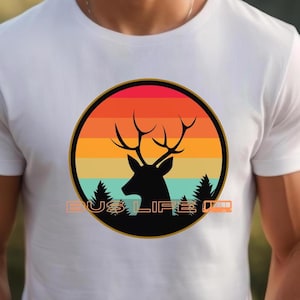 May include: White t-shirt with a circular design showing a deer silhouette against an orange, yellow, and red sunset. The design includes the text "BUS LIFE". The shirt is made of a soft material.