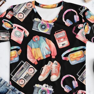 以下が含まれることがあります： 黒いVネックTシャツ。レトロなアイテムを水彩画でカラフルに描いたパターンです。デザインには、ラジカセ、カメラ、ヘッドフォン、ジャケット、スニーカー、電話が含まれています。シャツは、青いジーンズと黒いスニーカーと組み合わせて表示されています。