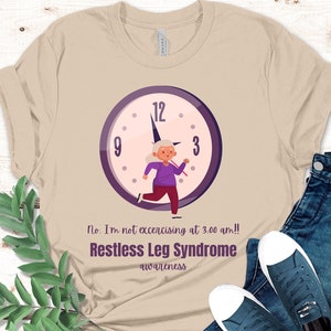 May include: A beige t-shirt featuring a purple clock face and a cartoon illustration of a person running. The text on the shirt reads "No. I'm not exercising at 3:00 am!! Restless Leg Syndrome awareness".