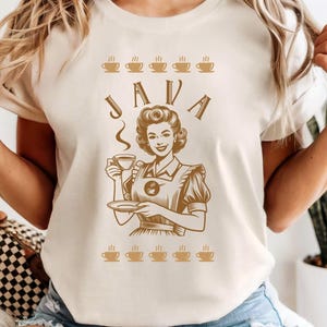 May include: Cream-coloured t-shirt featuring a vintage-style illustration of a woman holding a coffee cup, with the word "JAVA" above her. The design includes coffee cup illustrations. The shirt is short-sleeved.