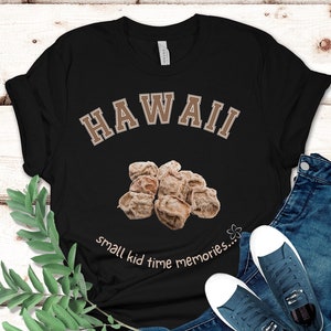 May include: A black short-sleeve t-shirt featuring the word HAWAII in a tan collegiate font, a graphic of several dried brown plums, and the text small kid time memories in a tan script font with a small flower icon next to the text.