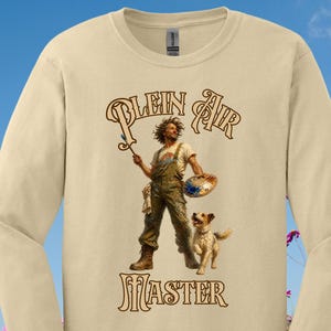 May include: A beige long-sleeve crewneck shirt featuring a graphic print of an artist in overalls holding a paintbrush and palette, accompanied by a small dog. The text "PLEIN AIR" appears above the figure and "MASTER" below in brown.