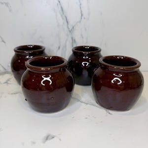 May include: Four small, round, dark brown ceramic pots with a glossy finish. Each pot has a wide opening and a slightly rounded body. The pots are arranged closely together, with a marble-patterned background.