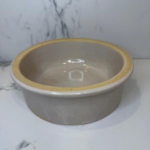 May include: A round, off-white ceramic bowl with a light yellow rim. The bowl has a simple, classic design and appears to be handmade. The interior is smooth and the exterior has a slightly textured surface.