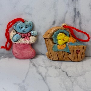 May include: Two vintage ceramic Christmas ornaments. One is a blue teddy bear wearing a pink stocking and a red bow. The other is a yellow chick in a brown wooden crib with a red heart.