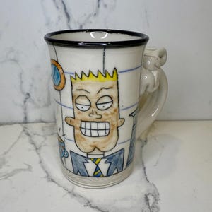 May include: White ceramic mug with a black rim and a handle. The mug features a cartoon illustration of a man with yellow hair, a blue suit, and a yellow tie. The mug has a unique handle design.