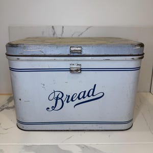 May include: Vintage white bread box with a hinged lid and a blue cursive "Bread" inscription. The box has blue stripes and metal clasps. A classic kitchen storage container.