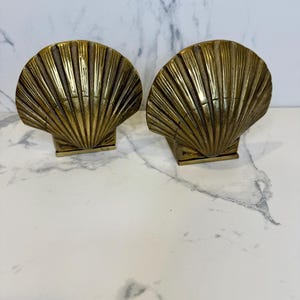 May include: A pair of brass bookends shaped like seashells. The bookends are a light gold color and have a detailed design.
