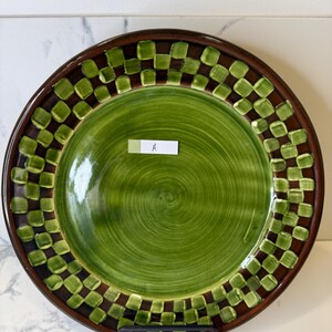 Op de afbeelding: Een ronde keramische bord met een effen groen midden en een zichtbaar wervelpatroon. De rand is versierd met een donkerbruin en lichtgroen geblokt patroon. Een klein wit rechthoekig label met de letter A is in het midden geplaatst.