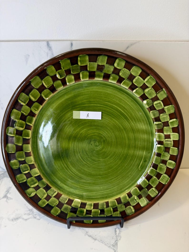 Op de afbeelding: Een ronde keramische bord met een effen groen midden en een zichtbaar wervelpatroon. De rand is versierd met een donkerbruin en lichtgroen geblokt patroon. Een klein wit rechthoekig label met de letter A is in het midden geplaatst.