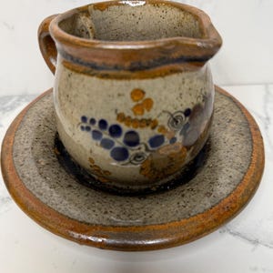 May include: A small, brown and white ceramic creamer with a handle and a floral design. It is sitting on a matching saucer.
