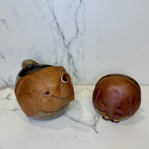 May include: Two ceramic elephant-shaped containers. One is a small pitcher with a handle and a brown and tan color scheme. The other is a small, round container with a lid. Both have smiling faces and are likely decorative or functional items.