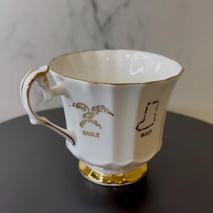 May include: White teacup with gold trim and a gold base. The cup features a gold eagle and boot design with the words "EAGLE" and "BOOT" printed below the images. The handle is also accented with gold.
