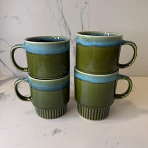 May include: Four ceramic mugs arranged in two stacks. Each mug features a deep olive green body with a contrasting light blue glazed rim. The bottom sections of the mugs have a vertical ribbed texture, while the upper portions are smooth.