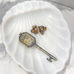 May include: A white, shell-shaped dish holds a vintage key pendant and a pair of clip-on earrings. The key pendant has a gold-colored frame with a decorative design and a gold-colored key. The earrings are gold-colored with amber-colored stones.
