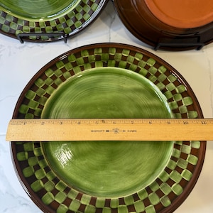 Op de afbeelding: Een rond keramieken bord met een groen midden en een donkerbruine en groene geblokte rand. Een houten liniaal ligt over het bord en toont een diameter van ongeveer 26,7 cm. Het bord heeft een gladde, glanzende afwerking.