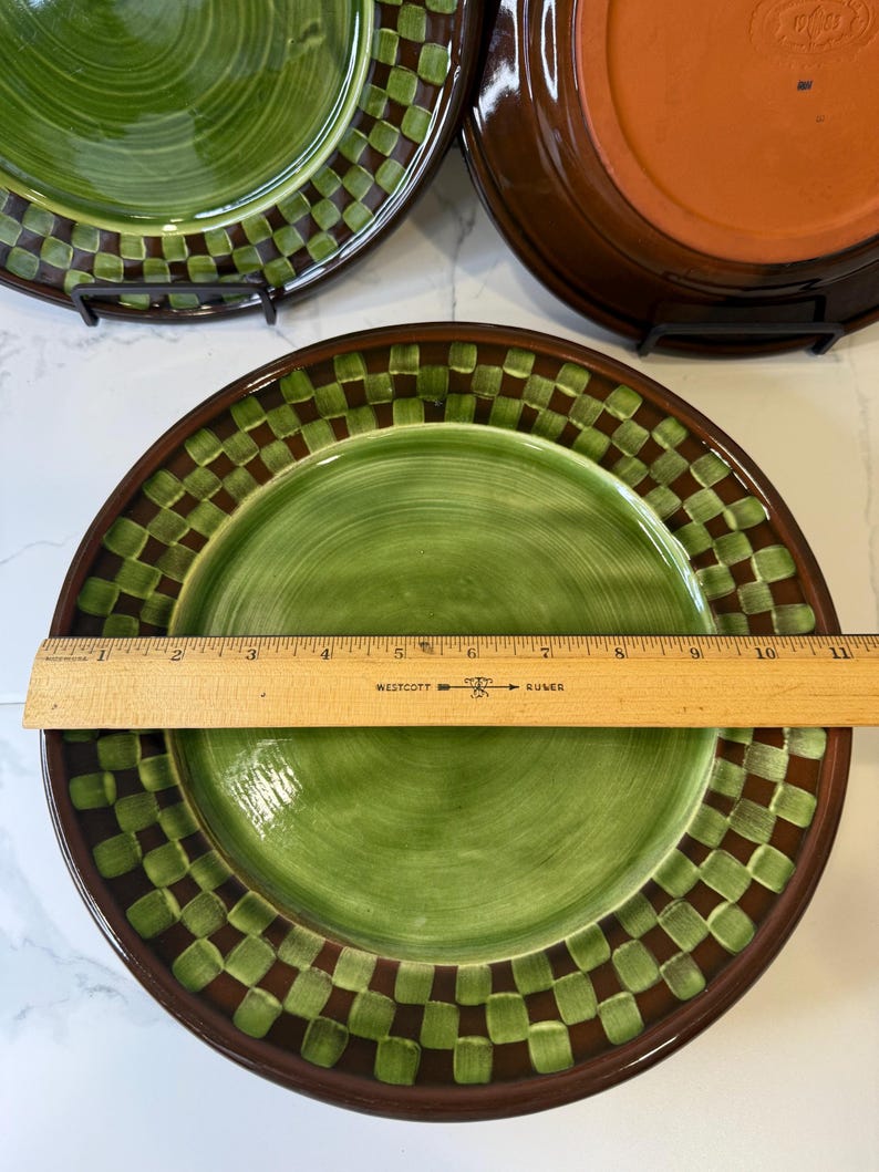 Op de afbeelding: Een rond keramieken bord met een groen midden en een donkerbruine en groene geblokte rand. Een houten liniaal ligt over het bord en toont een diameter van ongeveer 26,7 cm. Het bord heeft een gladde, glanzende afwerking.