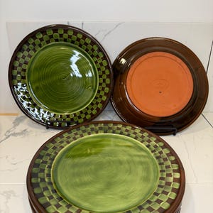 Op de afbeelding: Een set keramische borden met een groen cirkelvormig midden, omringd door een donkerbruine en groene geblokte rand. E&eacute;n bord staat ondersteboven en toont een effen terracotta-kleurige onderkant met een centraal in reli&euml;f aangebracht logo.