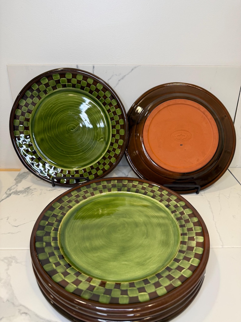 Op de afbeelding: Een set keramische borden met een groen cirkelvormig midden, omringd door een donkerbruine en groene geblokte rand. E&eacute;n bord staat ondersteboven en toont een effen terracotta-kleurige onderkant met een centraal in reli&euml;f aangebracht logo.