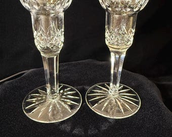 May include: A pair of clear glass candle holders featuring a faceted diamond-cut pattern on the upper bowl and a starburst design etched into the circular base. Each holder has a slender stem connecting the wide base to the flared top rim.