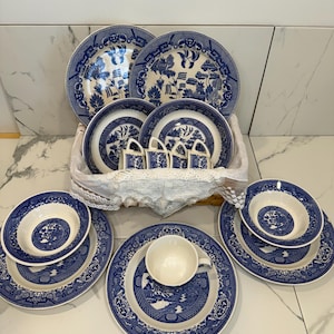 May include: A collection of blue and white porcelain dinnerware, including plates, bowls, and teacups. The set features a classic Willow pattern with detailed landscape scenes and decorative borders. The dishes are arranged in a basket and on a white surface.
