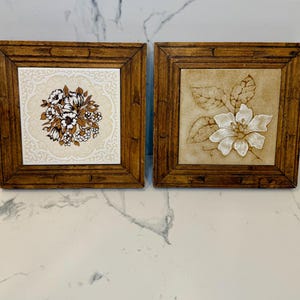May include: Two square decorative wall art pieces with wooden frames. One features a floral design in brown, white, and black on a white background. The other has a white flower with leaves on a beige background. Both are vintage style.