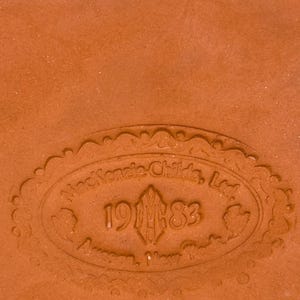 Op de afbeelding: Een terracotta-kleurig keramisch oppervlak met een in reli&euml;f aangebracht ovaal fabrieksmerk. De tekst luidt MacKenzie-Childs, Ltd. 1983 Aurora, New York, omringd door een decoratieve rand. Onder de ovale stempel staan de letters RW in zwarte inkt gedrukt.