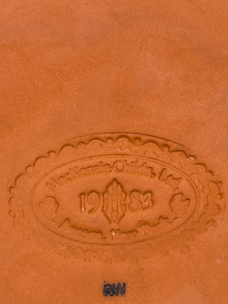 Op de afbeelding: Een terracotta-kleurig keramisch oppervlak met een in reli&euml;f aangebracht ovaal fabrieksmerk. De tekst luidt MacKenzie-Childs, Ltd. 1983 Aurora, New York, omringd door een decoratieve rand. Onder de ovale stempel staan de letters RW in zwarte inkt gedrukt.