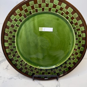 Op de afbeelding: Een rond keramieken bord met een effen groen midden en een brede rand versierd met een bruin en lichtgroen ruitpatroon. Een klein wit rechthoekig label met de handgeschreven letter E is in het midden van het bord geplaatst.