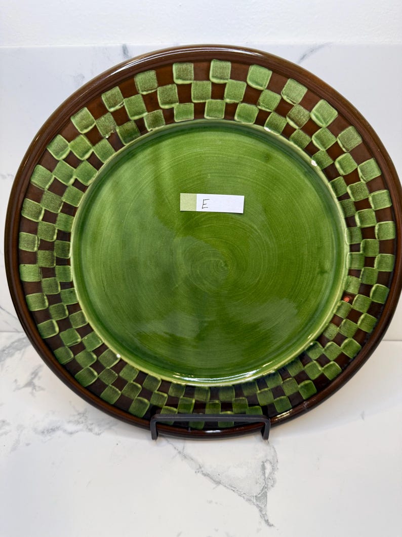 Op de afbeelding: Een rond keramieken bord met een effen groen midden en een brede rand versierd met een bruin en lichtgroen ruitpatroon. Een klein wit rechthoekig label met de handgeschreven letter E is in het midden van het bord geplaatst.