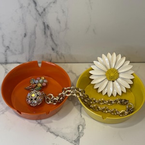 May include: An orange and yellow ashtray set with vintage jewelry. The orange ashtray holds a jeweled pendant and a floral brooch. The yellow ashtray holds a daisy brooch and a gold chain necklace. The background is a white marble.