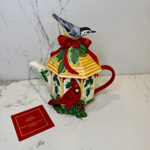 May include: A decorative teapot with a birdhouse design. The teapot features a yellow roof, red bow, and green holly accents. A red cardinal and a small bird are perched on the teapot. A red card with gold text reads "Winter Greetings Teapot".