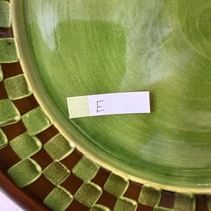 Op de afbeelding: Een ronde keramische bord met een glanzend limoengroen midden en een donkerbruine en groene geblokte rand. Een klein wit rechthoekig papieren label met de handgeschreven letter E is op het oppervlak van het bord geplaatst.
