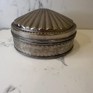 May include: A silver-colored, round jewelry box with a seashell-shaped lid. The box has a detailed, engraved band around the middle and a beaded trim. The box is sitting on a white marble surface.