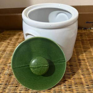 May include: A white ceramic canister with a cylindrical body and a matching lid. The lid features a deep green glaze with a central circular knob handle. The canister has a smooth, glossy finish and a wide opening at the top for storage.