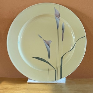 May include: A round, pale yellow ceramic plate with a floral design. The design features three calla lily blooms in shades of pink and gray, with green stems and leaves. The plate is resting on a white cloth.