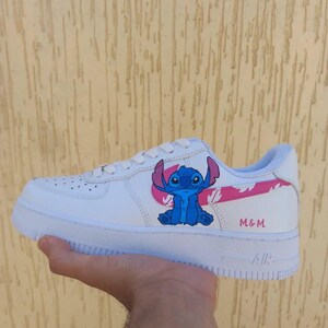 May include: White sneaker with a blue cartoon character design and pink accents. The sneaker features the text "M & M" and the word "AIR" on the sole. The design is on the side of the shoe.
