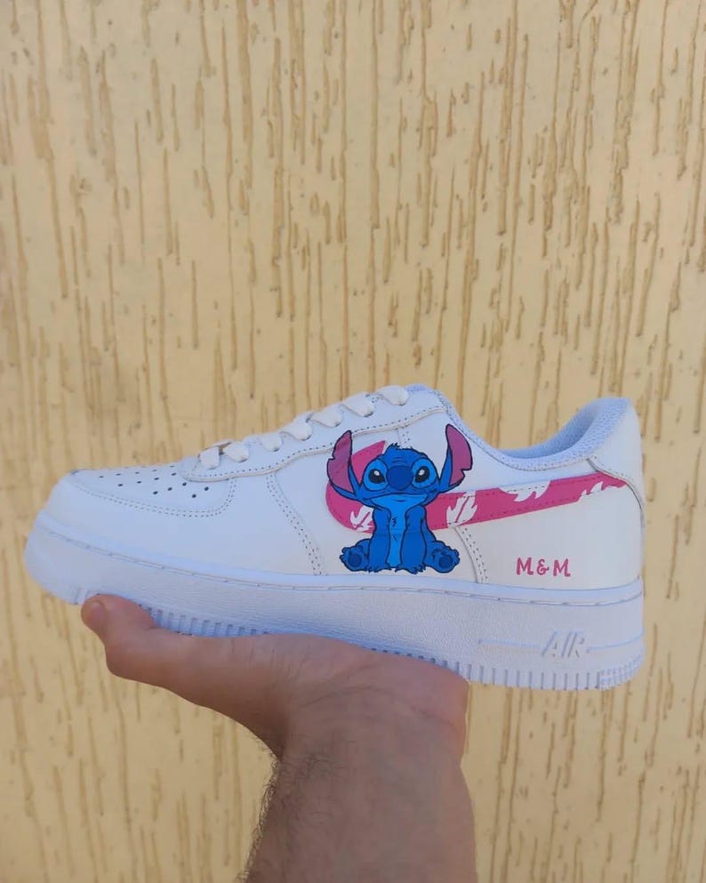 May include: White sneaker with a blue cartoon character design and pink accents. The sneaker features the text "M & M" and the word "AIR" on the sole. The design is on the side of the shoe.