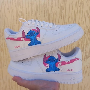 May include: White sneakers featuring a blue cartoon character with large ears and a pink accent. The text "M&M" is printed in pink on the side of the shoe. The shoes have white soles and laces.